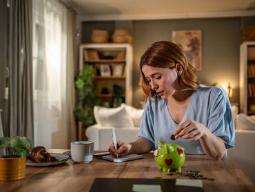 Young woman carefully managing her home finances, putting coins in a piggy bank and taking notes on expenses and savings in her living room