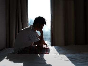 Side view of a depressed teenager boy sitting alone on a bed in bedroom