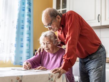 A pair of seniors working together at the kitchen table, focused on solving a crossword puzzle. While the woman sits and writes down the answers with a pencil, the man leans over with his hand on her shoulder and helps her with the answers, which adds a sense of mutual harmony and comfort to the whole moment.